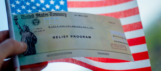 COVID-19 economic Stimulus check on blurred USA flag background. Relief program concept. Wide photo.