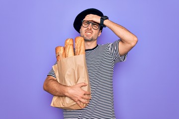 Young handsome baker man with blue eyes wearing french beret holding bag with bread stressed with hand on head, shocked with shame and surprise face, angry and frustrated. Fear and upset for mistake.