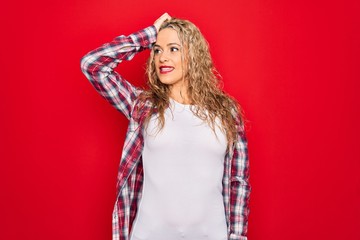 Young beautiful blonde woman wearing casual shirt standing over isolated red background smiling confident touching hair with hand up gesture, posing attractive and fashionable