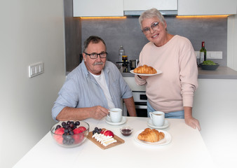 Senior couple having breakfast in the kitchen. Elderly man and woman enjoing  hot coffee or tea with fresh pastry croissant, jam, soft cheese and fruits. 