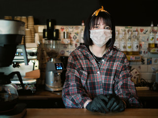Concept of biohazard security and social responsibility in coronavirus epidemic measures to contain virus spread: young barista girl in medical mask and medical gloves with city cafe background