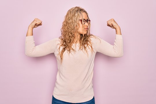 Young beautiful blonde woman wearing casual sweater and glasses over pink background showing arms muscles smiling proud. Fitness concept.