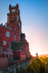 wall of a red castle on a hill with a tower and a clock against the sunset
