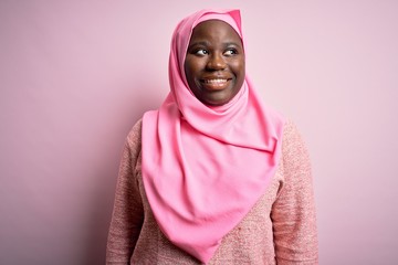 Young african american plus size woman wearing muslim hijab over isolated pink background looking away to side with smile on face, natural expression. Laughing confident.