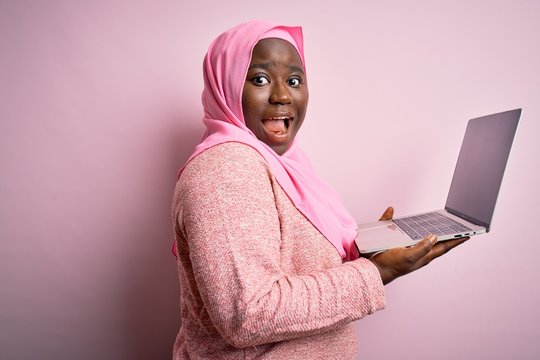African american plus size woman wearing muslim hijab using laptop over pink background pointing aside with hands open palms showing copy space, presenting advertisement smiling excited happy