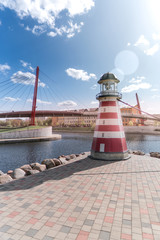 small striped lighthouse on the river bank on the background of the bridge and houses, focus on the lighthouse