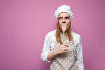 beautiful surprised girl cook in kitchen clothes with a spoon closes her mouth on a pink background, woman housewife with kitchen items