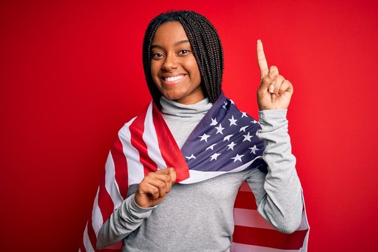 Young african american patriotic woman wearing united states of america flag on independence day surprised with an idea or question pointing finger with happy face, number one - Powered by Adobe