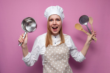 surprised girl cook in apron holds dishes and kitchen items on a pink background, portrait of a housewife woman