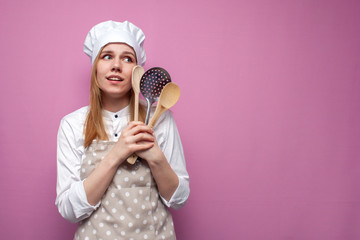 young cheerful girl cook in apron holds spoons and kitchen items and dreams on a pink background,...
