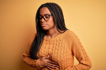 Young african american smart woman wearing glasses and casual sweater over yellow background with hand on stomach because indigestion, painful illness feeling unwell. Ache concept.