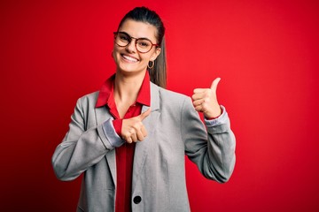 Young beautiful brunette businesswoman wearing jacket and glasses over red background Pointing to the back behind with hand and thumbs up, smiling confident