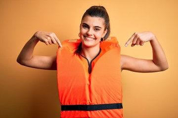Young beautiful brunette woman wearing orange safe lifejacket over yellow background looking...