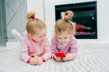 Two young focused children playing a smartphone with no name lying on the living room floor. Young children and technology, sisters play with a mobile phone, watch videos or play games