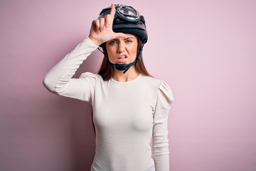 Young beautiful motorcyclist woman with blue eyes wearing moto helmet over pink background making fun of people with fingers on forehead doing loser gesture mocking and insulting.