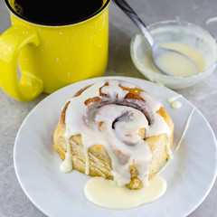 Cinnamon roll on a white plate with cup of coffee and bowl of glaze in the background.