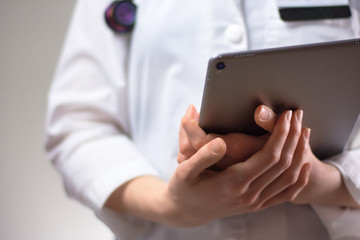 Tablet in the hands of healthcare professional up close. White coat, stethoscope, and badge visible in background. Hands of nurse practitioner or PA using technology in medicine for patients