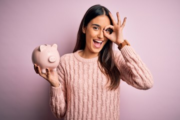 Young beautiful brunette woman holding piggy bank saving money for retirement with happy face smiling doing ok sign with hand on eye looking through fingers - obrazy, fototapety, plakaty