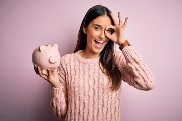 Fototapeten Young beautiful brunette woman holding piggy bank saving money for retirement with happy face smiling doing ok sign with hand on eye looking through fingers © Krakenimages.com