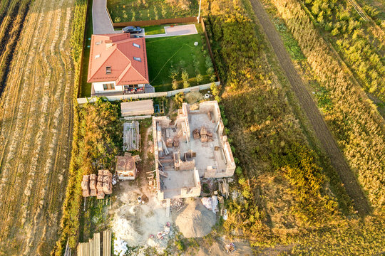 Top down aerial view of two private houses, one under construction with concrete foundament and brick walls and another finished with red tiled roof.