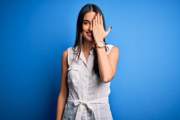 Young beautiful brunette woman wearing casual dress over isolated blue background covering one eye with hand, confident smile on face and surprise emotion.