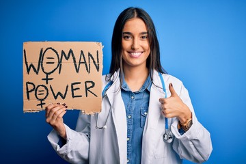 Young doctor woman wearing stethoscope holding cardboard banner with powe message happy with big smile doing ok sign, thumb up with fingers, excellent sign