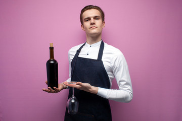 young guy sommelier holds a bottle of red wine on a colored background, an expert on wine in a white shirt and apron, the waiter offers wine