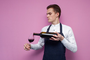 young professional waiter in uniform pours wine into a glass on a pink background, a sommelier guy tasting wine