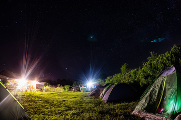 Night camping under starry sky. Glowing tourist tent, under beautiful evening sky full of stars and Milky way, city lights on background. Outdoor lifestyle concept