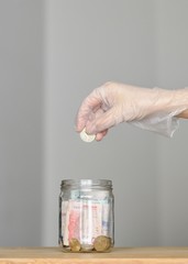 Mano con guante quirúrgico depositando dinero en un jarro de vidrio, ahorro de dinero durante la cuarentena - obrazy, fototapety, plakaty