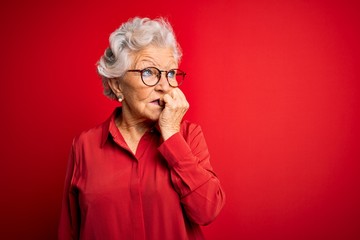 Senior beautiful grey-haired woman wearing casual shirt and glasses over red background looking...