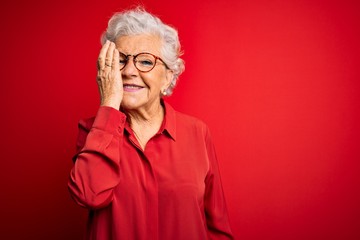 Senior beautiful grey-haired woman wearing casual shirt and glasses over red background covering one eye with hand, confident smile on face and surprise emotion.