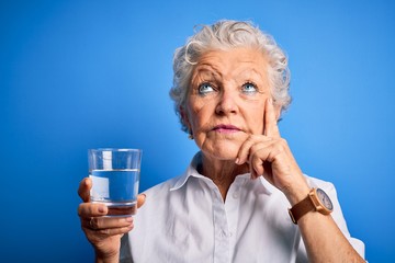 Senior beautiful woman drinking glass of water standing over isolated blue background serious face thinking about question, very confused idea