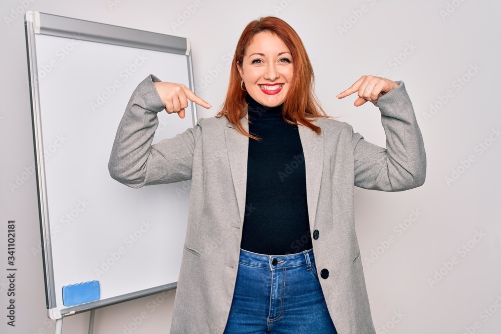 Wall mural Young beautiful redhead businesswoman doing business presentation using magnetic board looking confident with smile on face, pointing oneself with fingers proud and happy.