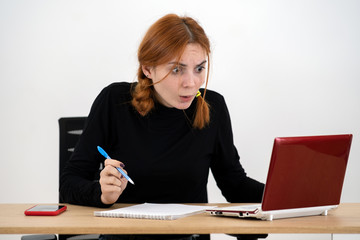 Shocked serious young office worker woman sitting behind working desk with laptop computer, cell phone and notebook.