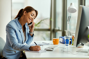 Happy businesswoman writing notes while talking on the phone in the office.