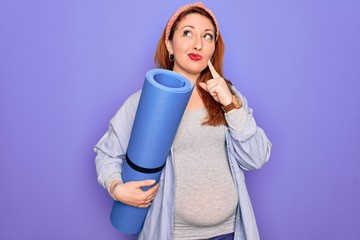 Young beautiful redhead pregnant woman expecting baby holding mat to do prenatal exercise serious face thinking about question, very confused idea