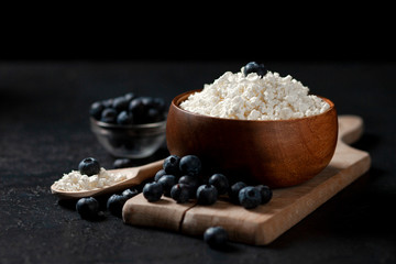 fresh homemade cottage cheese with blueberries in a wooden bowl against a dark background, healthy food with berries on a black table