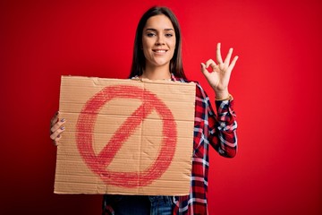 Young beautiful brunette woman holding banner with prohibited signal over red background doing ok...