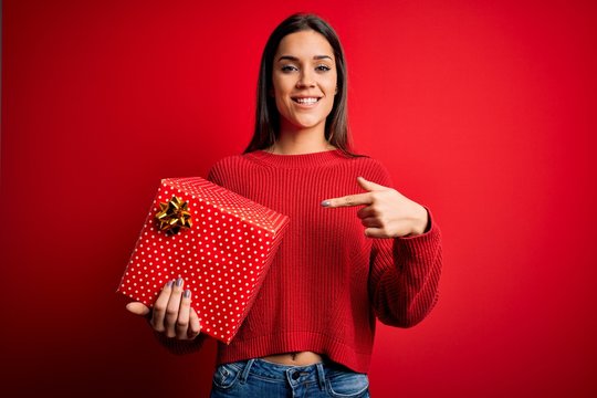Young beautiful brunette woman holding birthday gift over isolated red background very happy pointing with hand and finger
