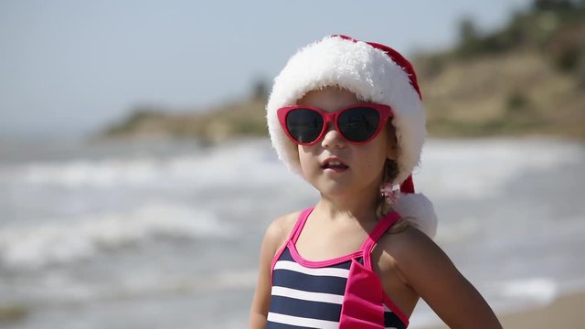 Little girl in Santa red hat having fun on tropical beach. Swim in the sea or ocean and points to the right. Christmas travel and vacation concept