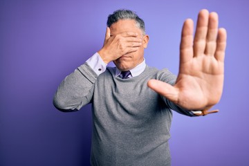 Middle age handsome grey-haired man wearing elegant sweater over purple background covering eyes...