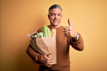 Middle age handsome grey-haired man holding paper bag with food over yellow background happy with big smile doing ok sign, thumb up with fingers, excellent sign