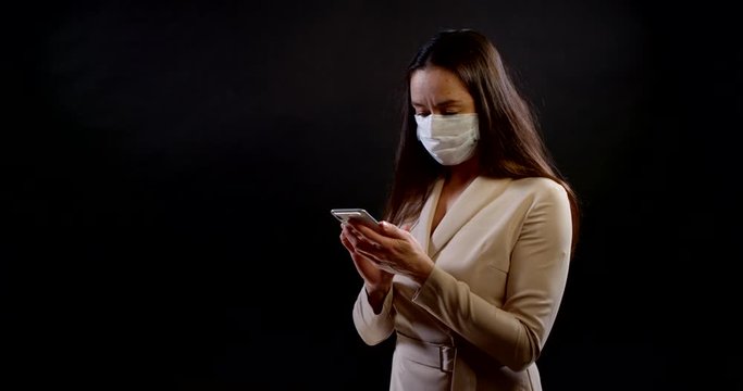 portrait of a woman on a black background. she's wearing a disposable face mask, standing three-quarters of the way up, holding her smartphone, looking at the screen, typing, and thinking