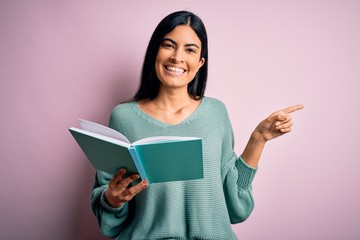Young beautiful hispanic student woman reading a book over pink isolated background very happy...