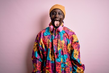 Obraz premium Young handsome african american man wearing colorful coat and cap over pink background sticking tongue out happy with funny expression. Emotion concept.