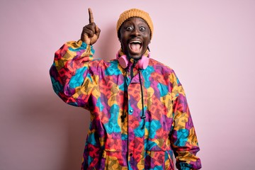 Young handsome african american man wearing colorful coat and cap over pink background pointing finger up with successful idea. Exited and happy. Number one.