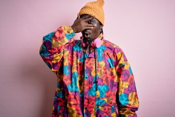 Young handsome african american man wearing colorful coat and cap over pink background peeking in...