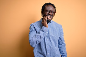 Fototapeta premium Young handsome african american man wearing shirt and glasses over yellow background touching mouth with hand with painful expression because of toothache or dental illness on teeth. Dentist