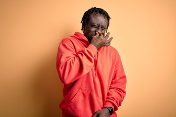 Young african american sporty man wearing sweatshirt with hoodie over yellow background smelling something stinky and disgusting, intolerable smell, holding breath with fingers on nose. Bad smell